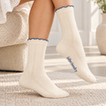A person seated indoors wearing cream-colored ribbed crew socks with blue scalloped trim and blue 'baby laundry' text near the cuff, shown on a textured rug in a bright room.