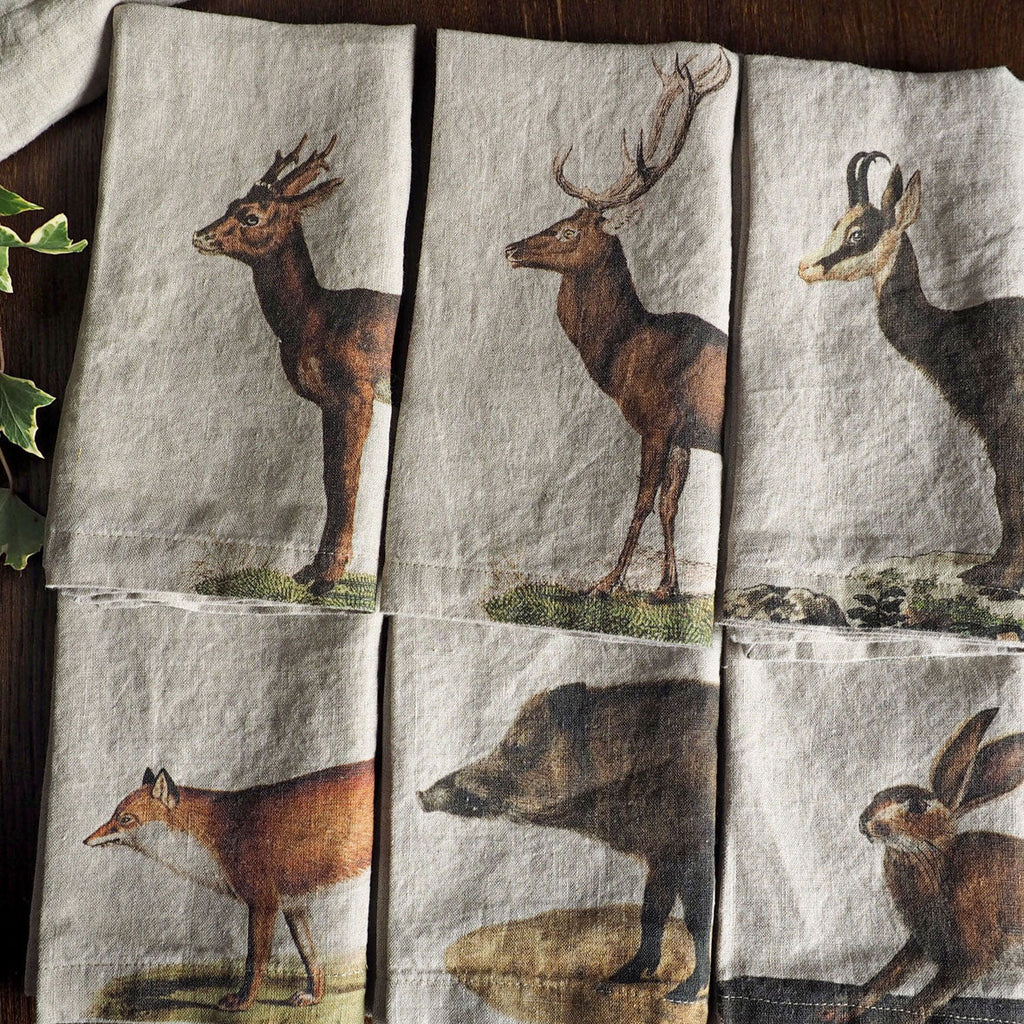 Six square linen napkins with a rustic natural beige background, each printed with a different woodland animal illustration (deer, fox, hare, hyena, bird, and another deer). The napkins are arranged in a 2x3 grid on a dark wooden table.