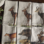 Six square linen napkins with a rustic natural beige background, each printed with a different woodland animal illustration (deer, fox, hare, hyena, bird, and another deer). The napkins are arranged in a 2x3 grid on a dark wooden table.