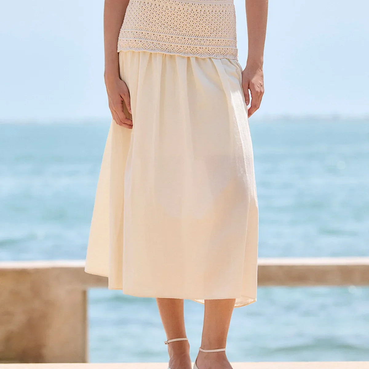 Woman wearing a light yellow dress with a textured waistband, standing by a pool with water in the background.