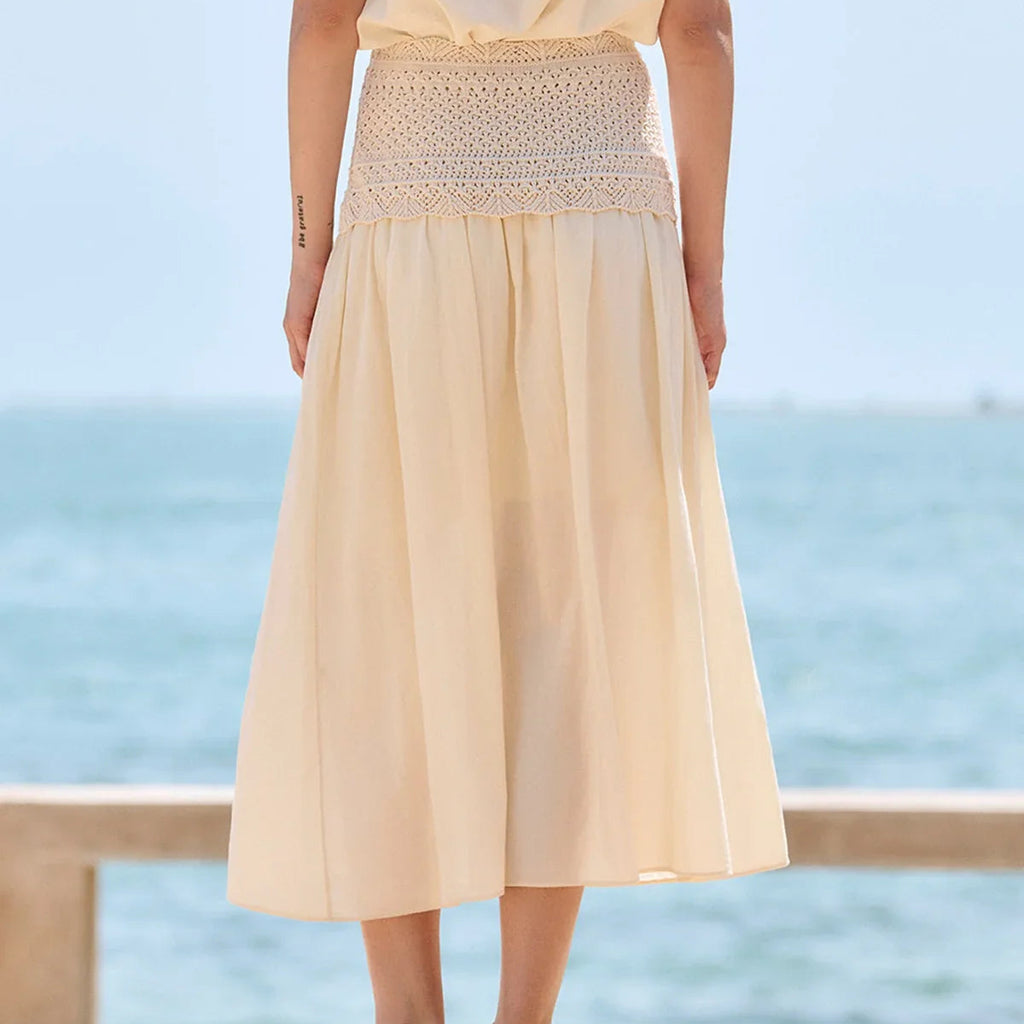 A woman wearing a light beige midi skirt with a smocked crochet waistband, standing outdoors with the ocean in the background.