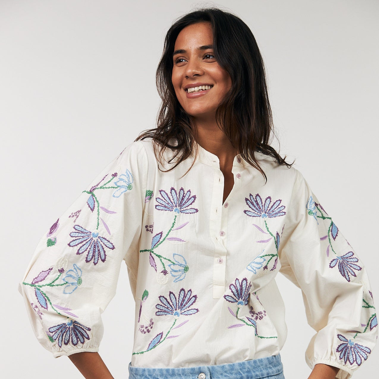 Woman wearing a white blouse with floral patterns and blue jeans against a white background