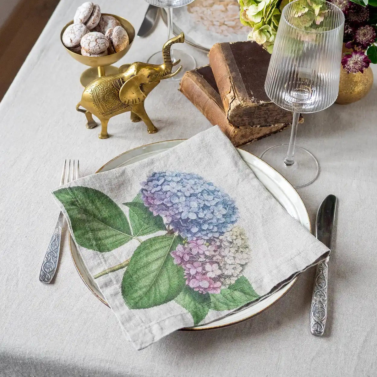 A styled table setting featuring a linen napkin with a watercolor-style print of purple and blue hydrangeas with green leaves, folded on a gold-rimmed plate. The setting includes a silver knife, fork, ribbed wine glasses, a small gold elephant figurine, and a floral centerpiece on a light beige tablecloth.