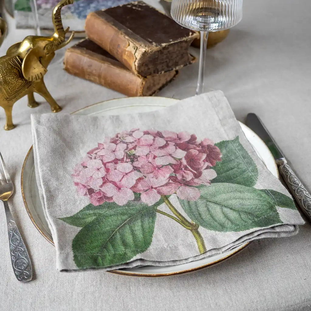A folded linen napkin with a detailed print of a pink hydrangea and green leaves, styled on a table setting with a white plate, silver cutlery, a gold elephant figurine, and vintage books.