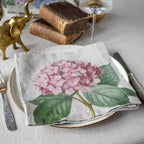 A folded linen napkin with a detailed print of a pink hydrangea and green leaves, styled on a table setting with a white plate, silver cutlery, a gold elephant figurine, and vintage books.