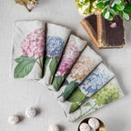 A set of six folded linen napkins with a watercolor-style hydrangea floral print, arranged in a fan shape on a light beige linen surface. The napkins feature various hydrangea colors including pink, blue, peach, and green on a light gray-beige linen base.
