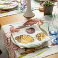 A styled dining table setting featuring a linen table runner and matching napkins with a colorful seashell and fish print, paired with white plates, cutlery, and glassware on a light wood table.