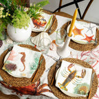 A styled outdoor dining table setting with a white tablecloth printed with red and green sea life motifs, woven round placemats, white plates, matching napkins featuring crab, lobster, seahorse, and starfish prints, a white vase of greenery, and a yellow taper candle.