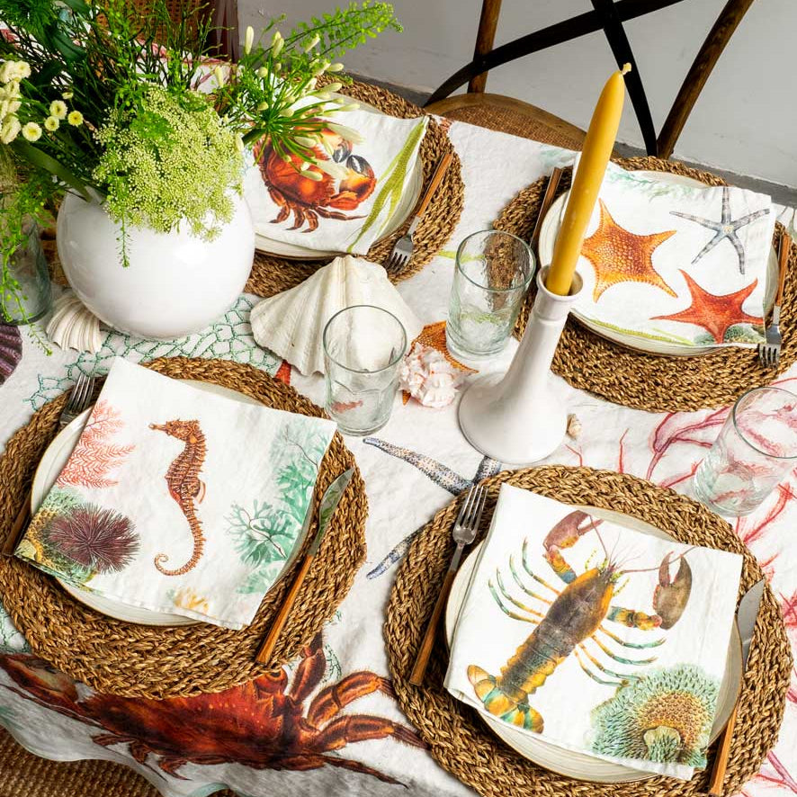 A styled outdoor dining table setting with a white tablecloth printed with red and green sea life motifs, woven round placemats, white plates, matching napkins featuring crab, lobster, seahorse, and starfish prints, a white vase of greenery, and a yellow taper candle.