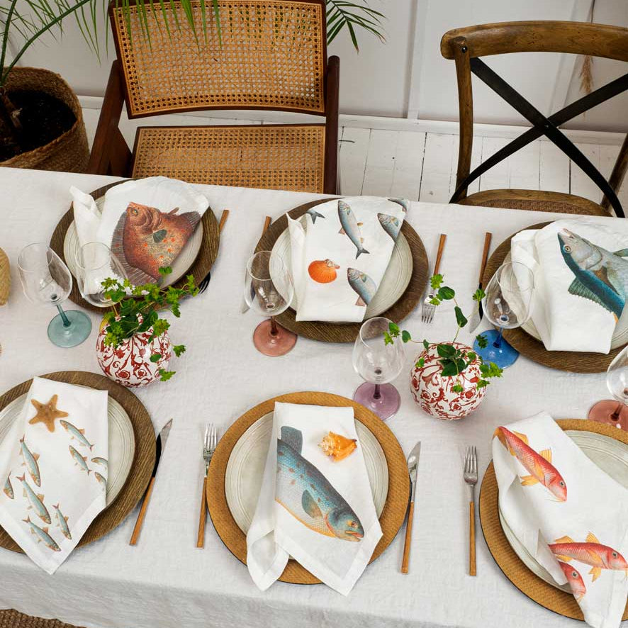 A styled dining table set with white tablecloths, round brown plates, and white napkins printed with colorful fish illustrations. The table is set with wine glasses, cutlery, small floral pots, and chairs in the background.