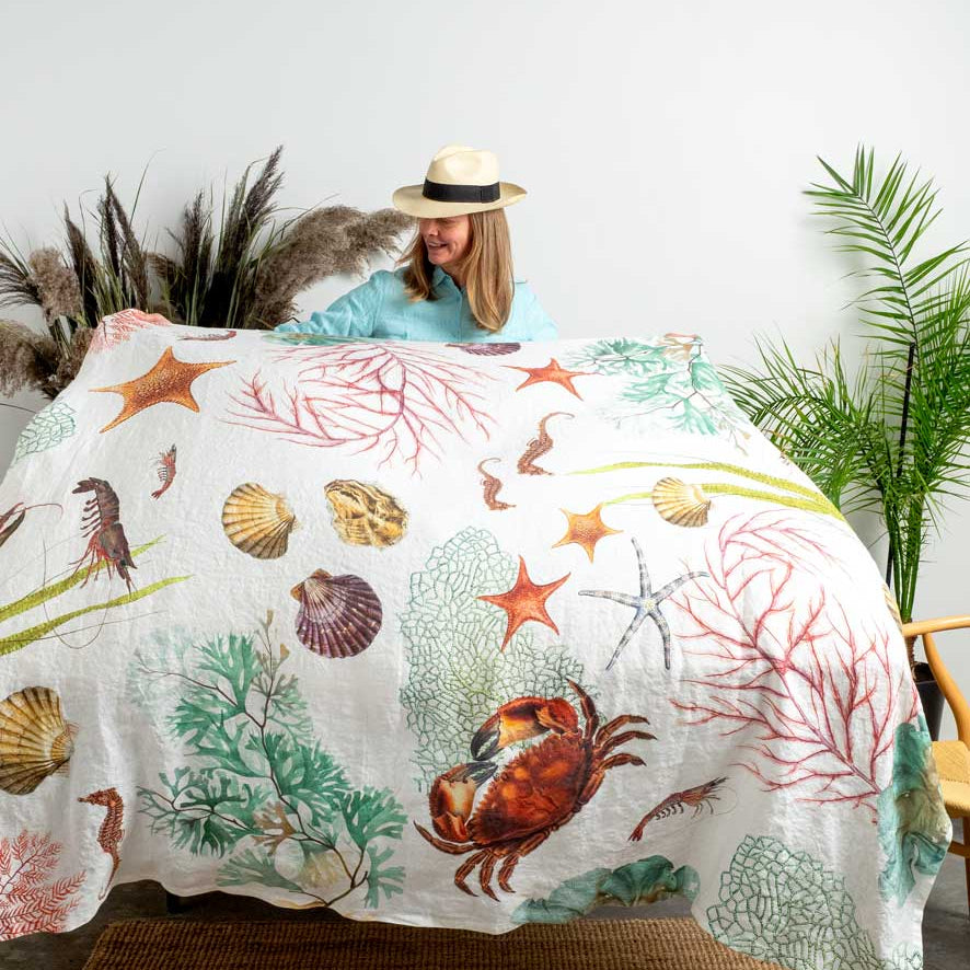 A woman wearing a straw hat and light blue top holds up a white quilt or bedspread printed with colorful sea creatures and coral, shown in a styled bedroom setting with plants and a woven rug.
