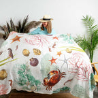 A woman wearing a straw hat and light blue top holds up a white quilt or bedspread printed with colorful sea creatures and coral, shown in a styled bedroom setting with plants and a woven rug.