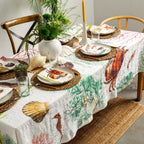 A white tablecloth with a colorful nautical print of crabs, seashells, coral, and sea plants, styled on a dining table with plates, woven placemats, and a white elephant vase with greenery.