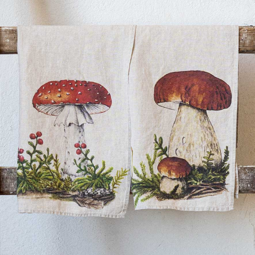 Two cream-colored linen kitchen towels hanging from a rustic wooden rack against a textured white wall. The left towel features an illustration of a red-and-white fly agaric mushroom with green foliage and red berries.
