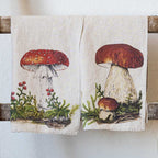Two cream-colored linen kitchen towels hanging from a rustic wooden rack against a textured white wall. The left towel features an illustration of a red-and-white fly agaric mushroom with green foliage and red berries.