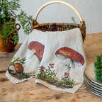 A woven wicker basket with a natural wood handle sits on a rustic wooden table, draped with a cream linen cloth printed with red and brown mushrooms and small red berries. A potted green plant and stacked plates are visible in the background.