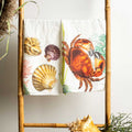 A white kitchen towel featuring a colorful print of a red-orange crab, seashells, and sea plants, draped over a bamboo ladder stand with decorative seashells on a woven tray below.