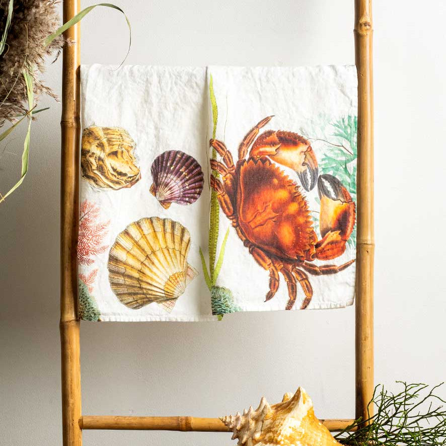 A white kitchen towel featuring a colorful print of a red-orange crab, seashells, and sea plants, draped over a bamboo ladder stand with decorative seashells on a woven tray below.
