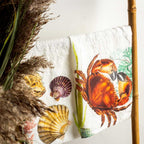 A white kitchen towel with a colorful seashell and orange crab print, draped over a bamboo stand next to dried pampas grass against a white wall.