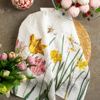 A white kitchen tea towel with a colorful watercolor print of tulips, daffodils, and a bee, styled in a flat-lay arrangement with pink tulips, a glass bowl of meringues, and a decorative ceramic pumpkin on a gray stone surface.