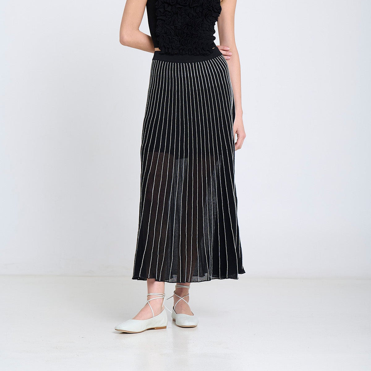 A model stands against a plain white studio background wearing a black sleeveless top with ruffled detailing and a long black pleated midi skirt with thin vertical white stripes, paired with white flat shoes.