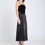 A woman stands in profile wearing a black sleeveless ruffled top and a long black pleated skirt with thin vertical white stripes, paired with light beige shoes, against a plain white studio background.