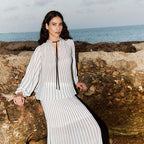 A woman wearing a white and gray vertically striped maxi dress with a tie-neck detail, posing on a rocky beach with the ocean in the background.