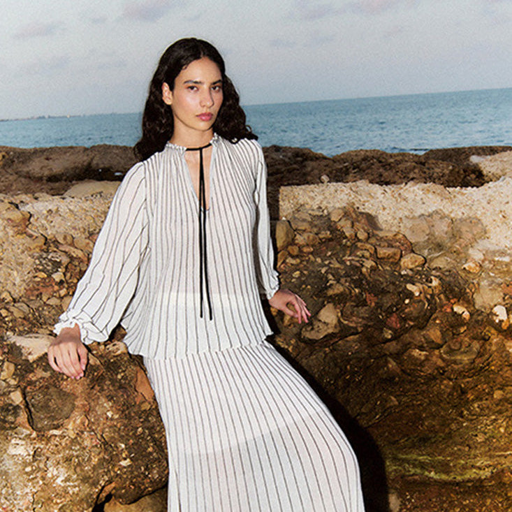 A woman wearing a white and gray vertically striped maxi dress with a tie-neck detail, posing on a rocky beach with the ocean in the background.