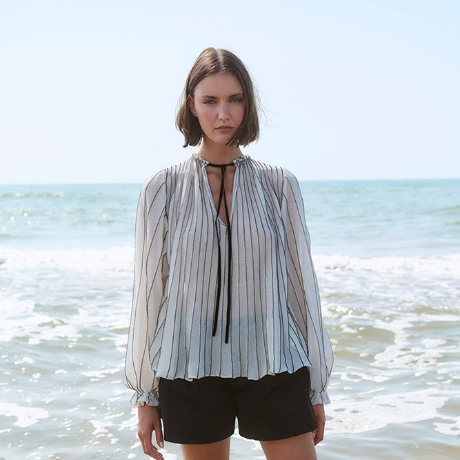 Woman standing in shallow water at the beach wearing a striped blouse and black shorts.