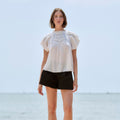 A woman stands outdoors by the water wearing a white short-sleeve blouse with puff sleeves and a decorative embroidered yoke, paired with black shorts and brown platform sandals.