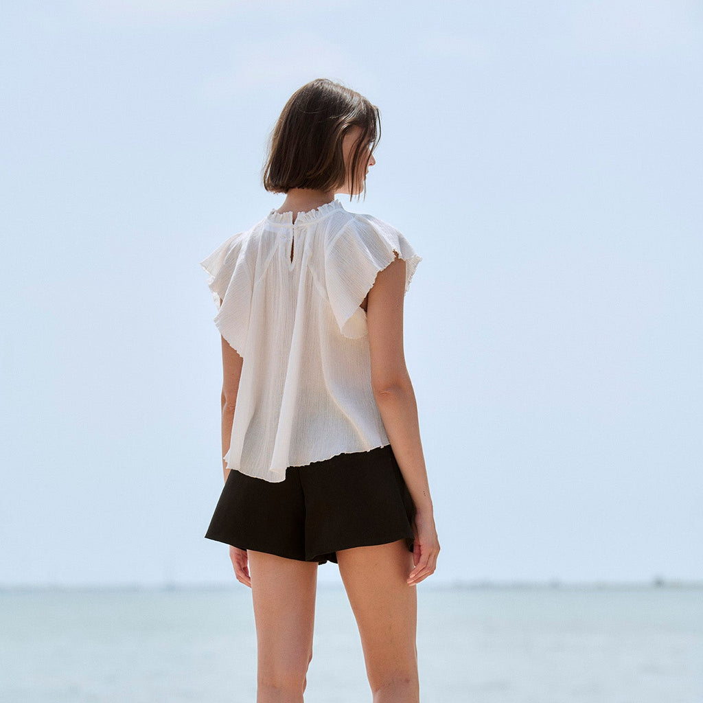 A woman stands with her back to the camera on a stone pier overlooking a calm sea under a pale sky, wearing a white ruffled sleeveless blouse and black shorts.
