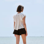 A woman stands with her back to the camera on a stone pier overlooking a calm sea under a pale sky, wearing a white ruffled sleeveless blouse and black shorts.