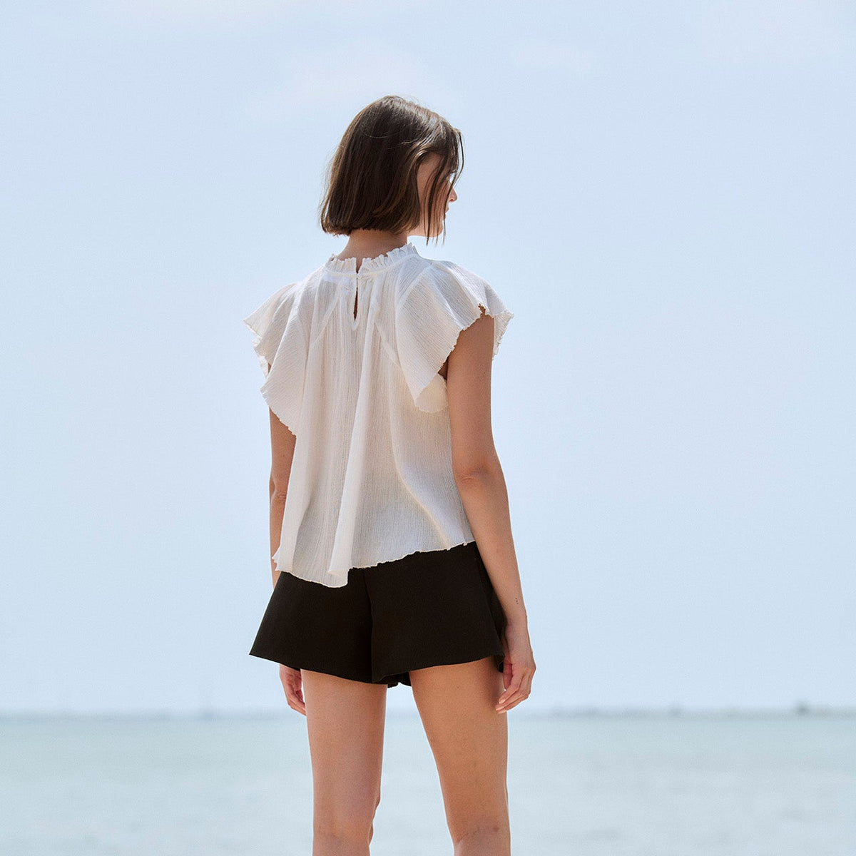 A woman stands with her back to the camera on a stone pier overlooking a calm sea under a pale sky, wearing a white ruffled sleeveless blouse and black shorts.
