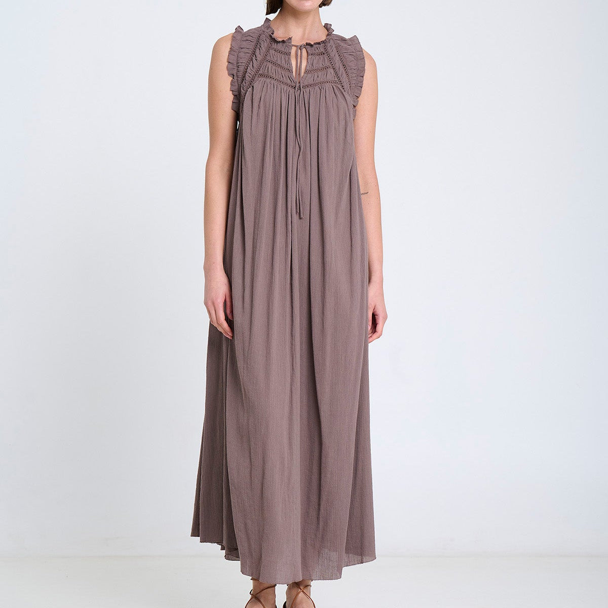 A woman modeling a sleeveless, floor-length taupe dress with ruffled shoulder details and a gathered neckline, standing against a plain white studio background.