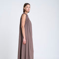 A woman modeling a sleeveless, floor-length taupe dress with a draped shawl collar and gathered neckline, paired with brown leather flat shoes, set against a plain white studio background.