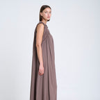 A woman modeling a sleeveless, floor-length taupe dress with a draped shawl collar and gathered neckline, paired with brown leather flat shoes, set against a plain white studio background.