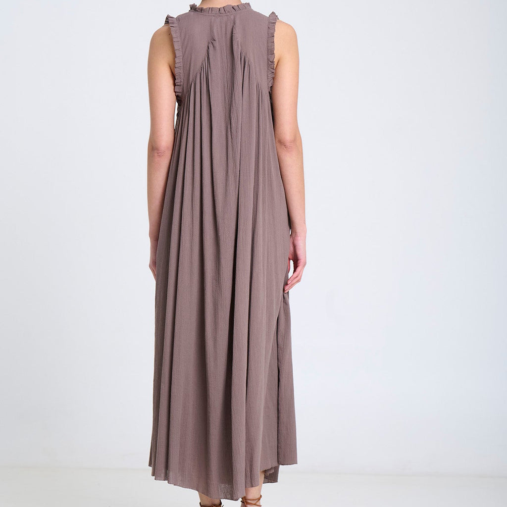 A woman viewed from behind wearing a sleeveless, floor-length pleated dress in a muted taupe-brown color, paired with brown strappy flat shoes, against a plain white studio background.