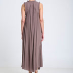A woman viewed from behind wearing a sleeveless, floor-length pleated dress in a muted taupe-brown color, paired with brown strappy flat shoes, against a plain white studio background.