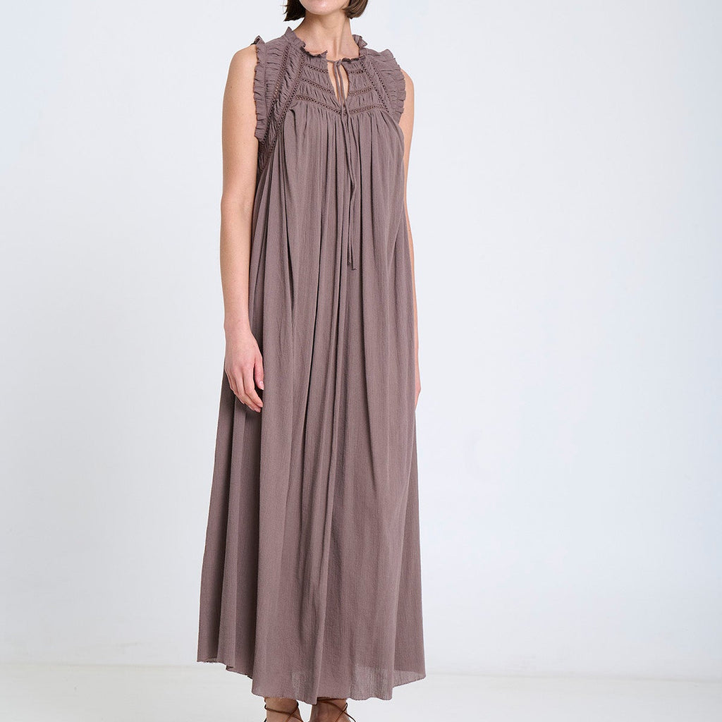 A woman modeling a sleeveless, floor-length taupe dress with a gathered neckline and tie detail, paired with brown flat shoes, set against a plain white studio background.