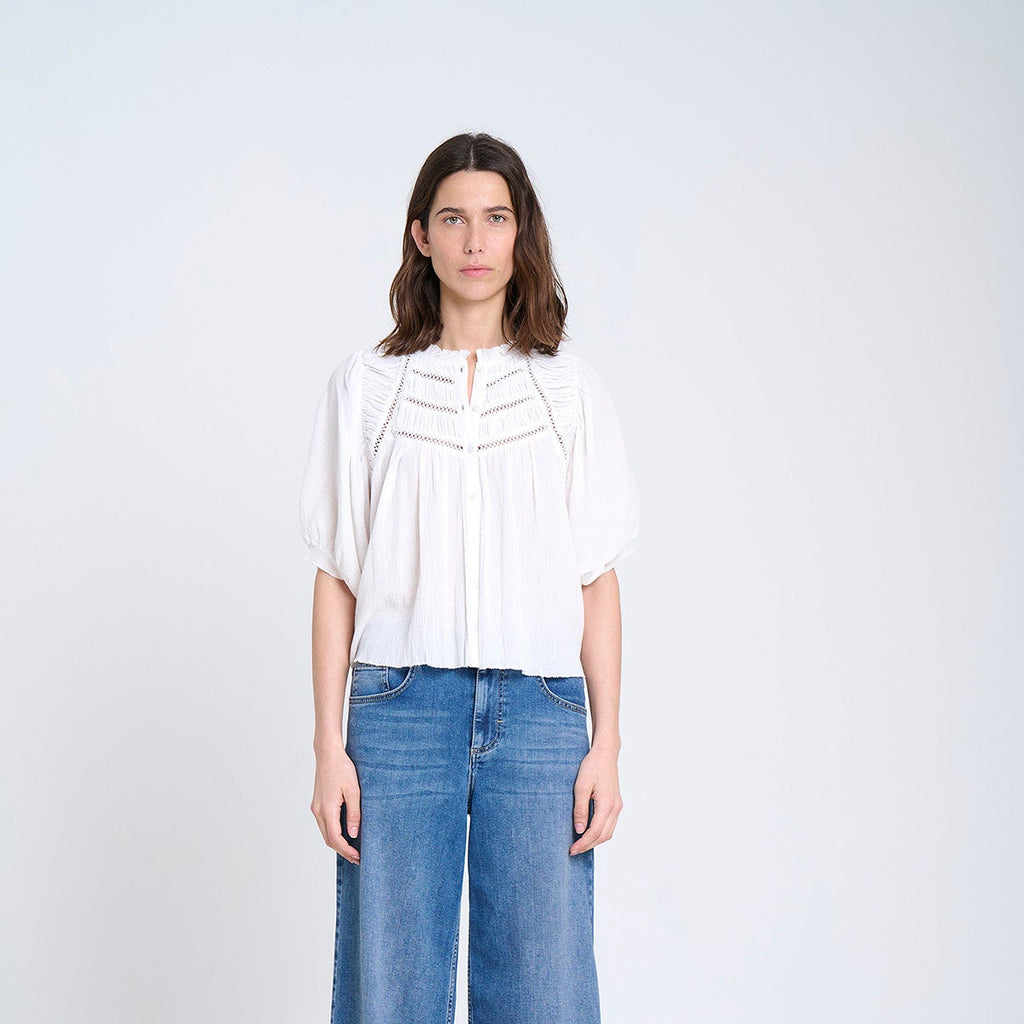 A woman stands against a plain white studio background wearing a white short-sleeve blouse with decorative chest detailing and wide-leg blue jeans.