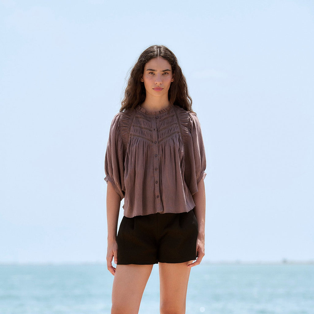 A woman stands outdoors by the sea wearing a loose taupe-brown short-sleeve blouse, black shorts, and white flat shoes.