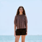 A woman stands outdoors by the sea wearing a loose taupe-brown short-sleeve blouse, black shorts, and white flat shoes.