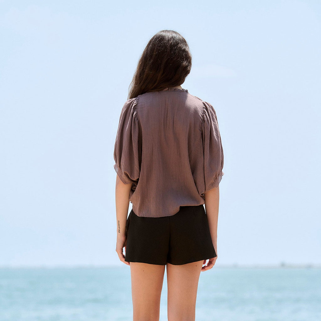 A woman viewed from behind stands on a sunny beachside walkway wearing a sheer taupe puff-sleeve blouse, black shorts, and white lace-up sandals, with the blue sea and sky in the background.