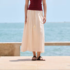 A woman stands outdoors by the sea wearing a sleeveless burgundy top and a long cream skirt, with sandals, against a blue sky and ocean background.