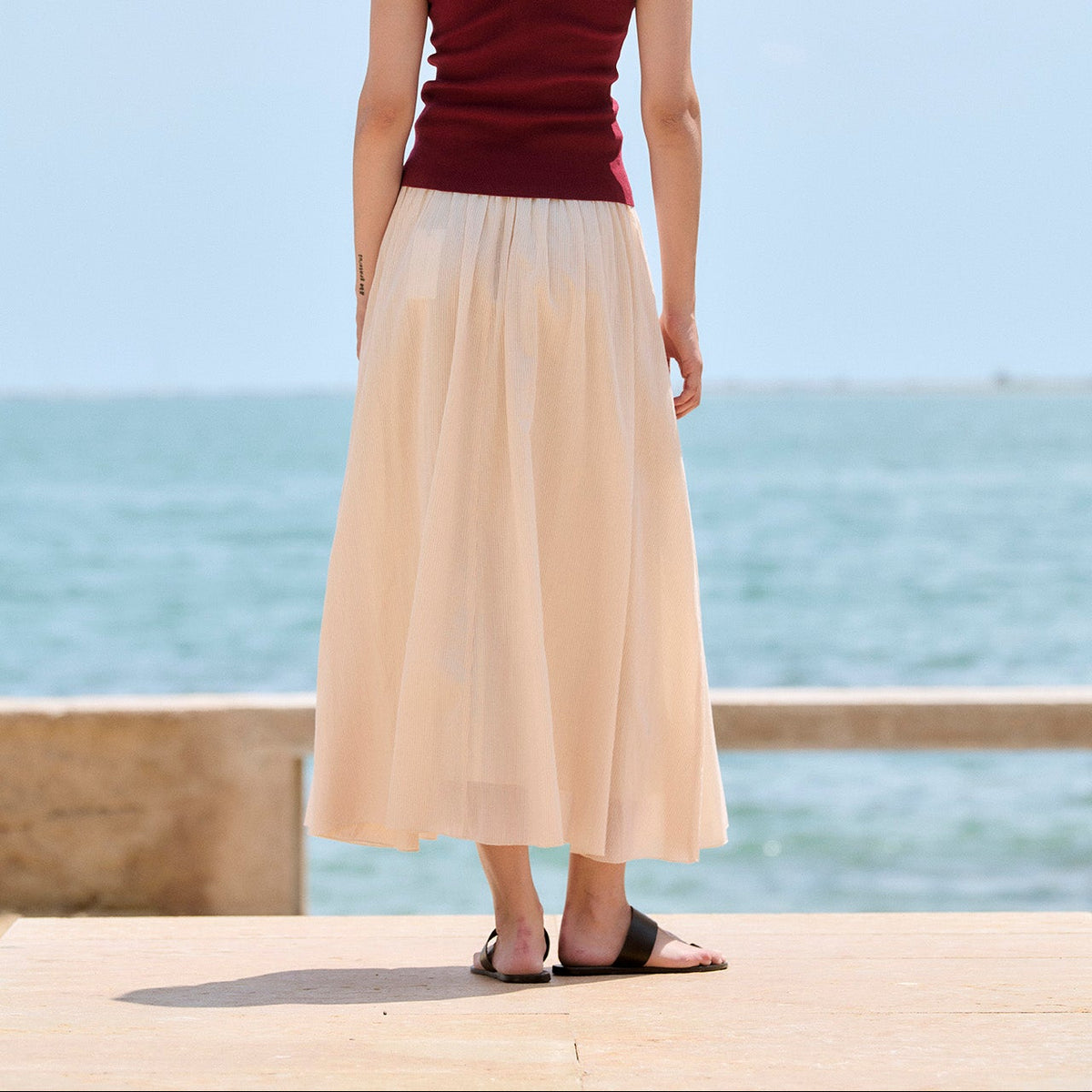 A woman stands with her back to the camera on a sunny day by the sea, wearing a dark red spaghetti-strap tank top and a long, flowing light beige maxi skirt.