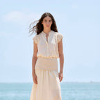 A woman stands outdoors by the sea wearing a light cream sleeveless blouse with lace cap sleeves and a matching midi skirt. The background shows a pale blue sky and ocean.