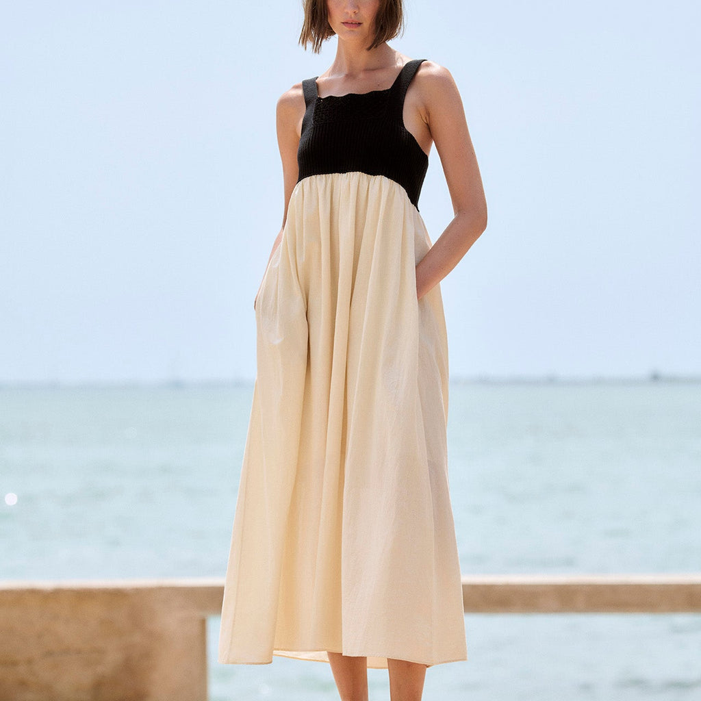 A woman stands outdoors by the sea wearing a sleeveless midi dress with a black bodice and a flowing cream skirt, paired with black sandals.