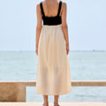 A woman stands with her back to the camera on a sunny day by the sea, wearing a sleeveless maxi dress with a black sweetheart-style bodice and a flowing cream-colored skirt.