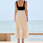 A woman stands with her back to the camera on a sunny day by the sea, wearing a sleeveless maxi dress with a black sweetheart-style bodice and a flowing cream-colored skirt.
