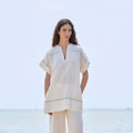A woman stands outdoors by the sea wearing a loose off-white linen two-piece set with short sleeves and wide-leg pants, featuring thin dark contrast stripes.